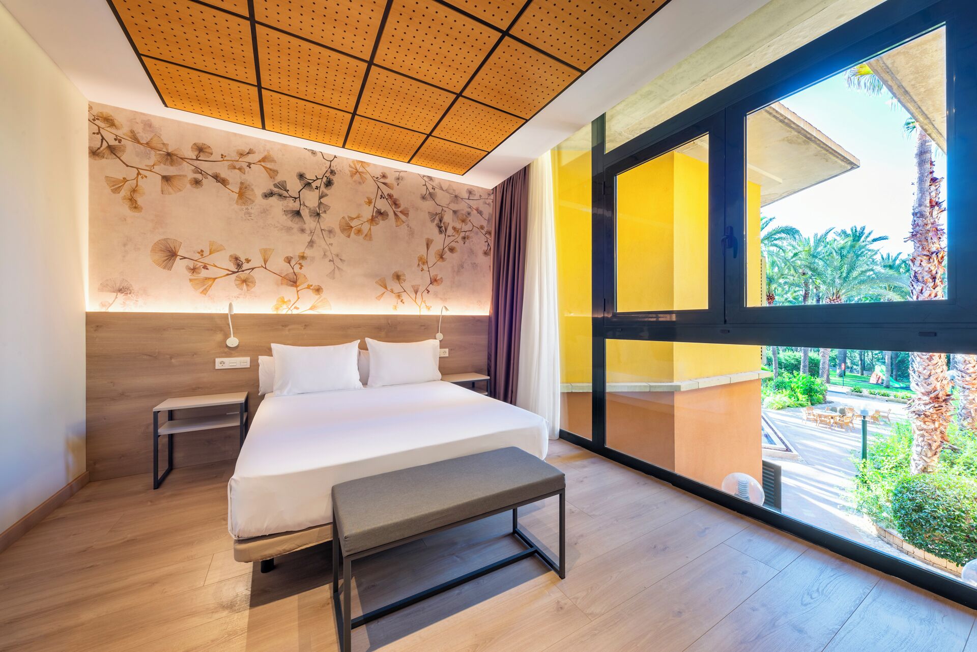 A modern hotel room with a large bed, a bedside table, a bench, a ceiling with a perforated design, and a view of a garden through large windows.