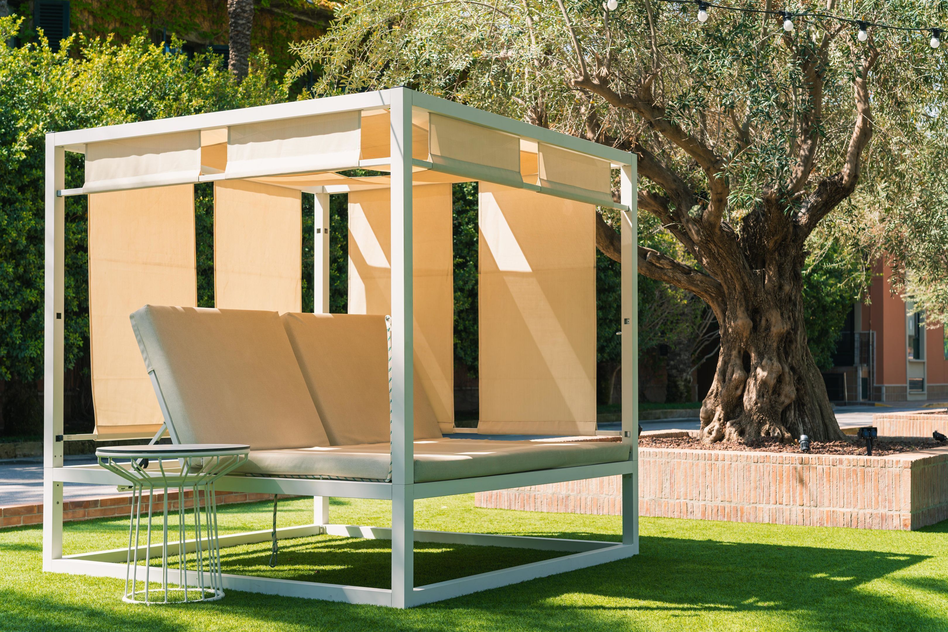 A modern outdoor seating area with a metal frame and beige cushions, surrounded by a well-maintained lawn and a large tree.