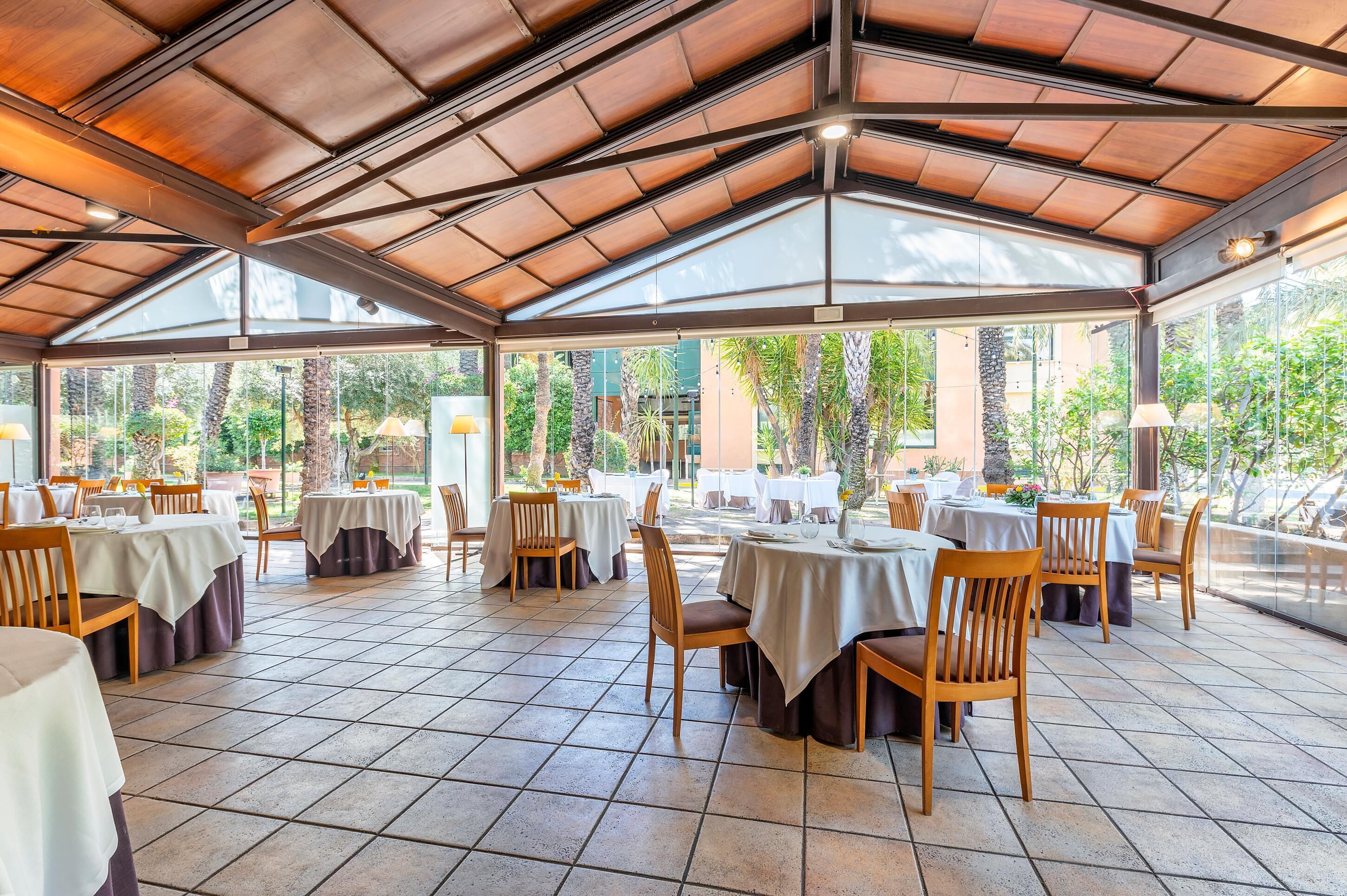 Un área de comedor al aire libre con techo, mesas y sillas, ventanas amplias y piso de baldosas.