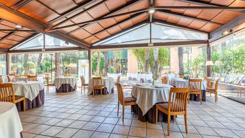 Un área de comedor al aire libre con techo, mesas y sillas, ventanas amplias y piso de baldosas.
