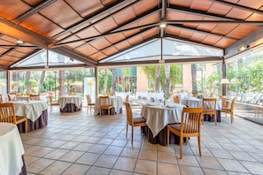 Un área de comedor al aire libre con techo, mesas y sillas, ventanas amplias y piso de baldosas.