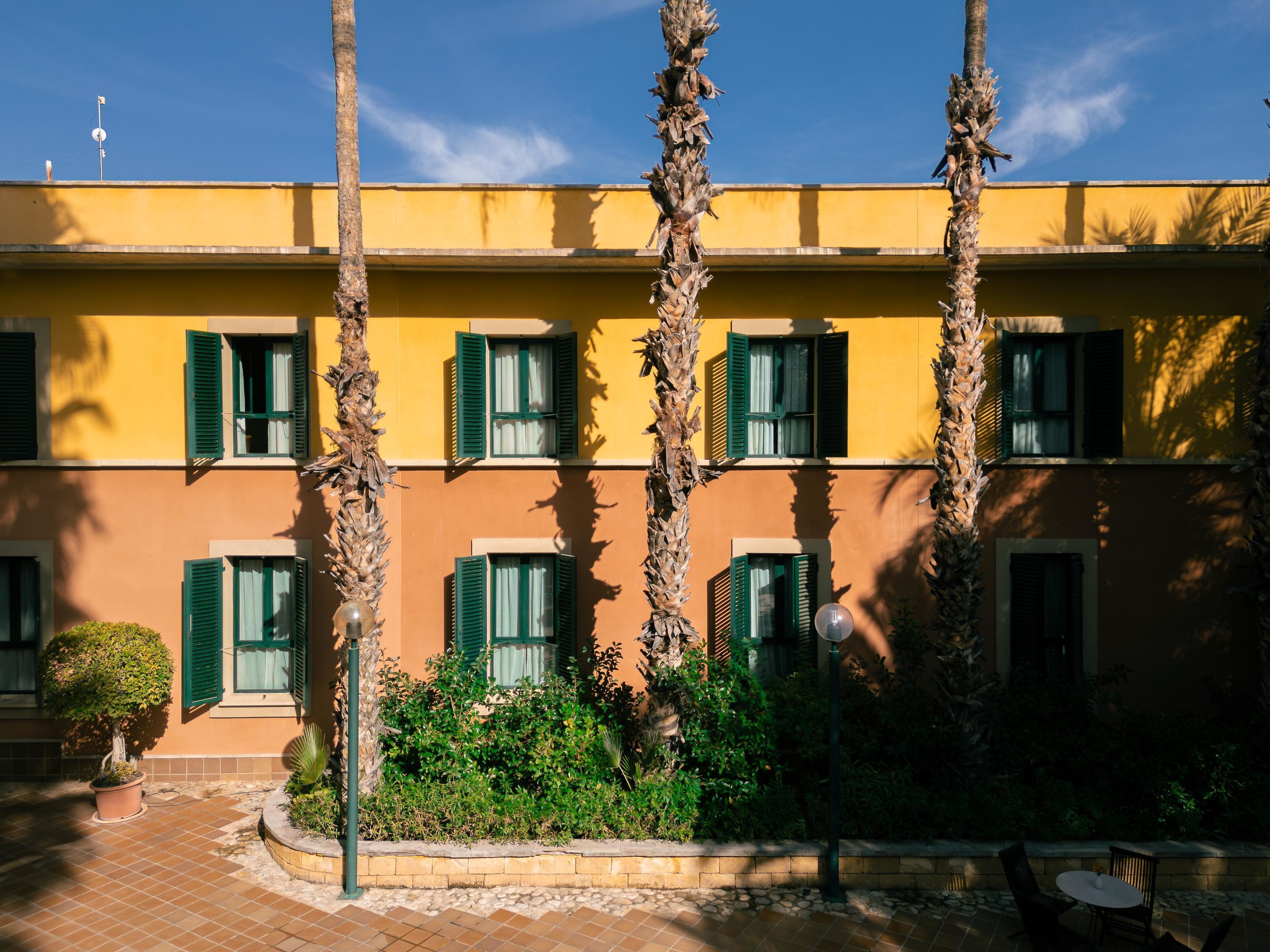 Un edificio con varias ventanas y contraventanas verdes, rodeado de palmeras y un patio con baldosas.