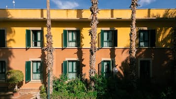 Un edificio con varias ventanas y contraventanas verdes, rodeado de palmeras y un patio con baldosas.