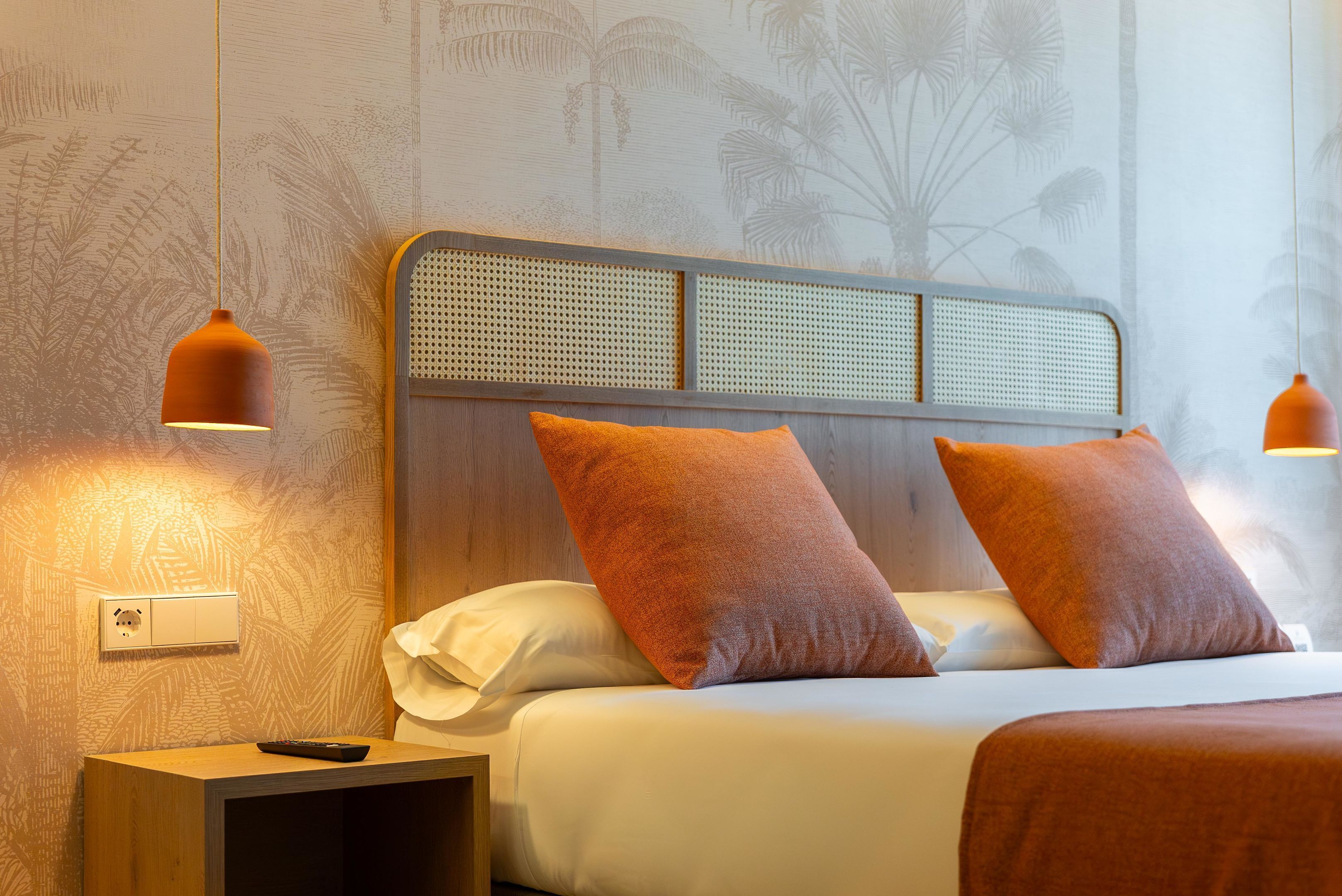 Una cama bien hecha con dos almohadas naranjas, una mesita de noche de madera con un control remoto y dos lámparas colgantes.