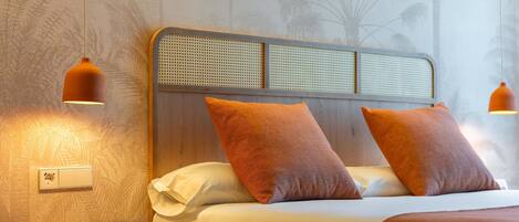 Una cama bien hecha con dos almohadas naranjas, una mesita de noche de madera con un control remoto y dos lámparas colgantes.