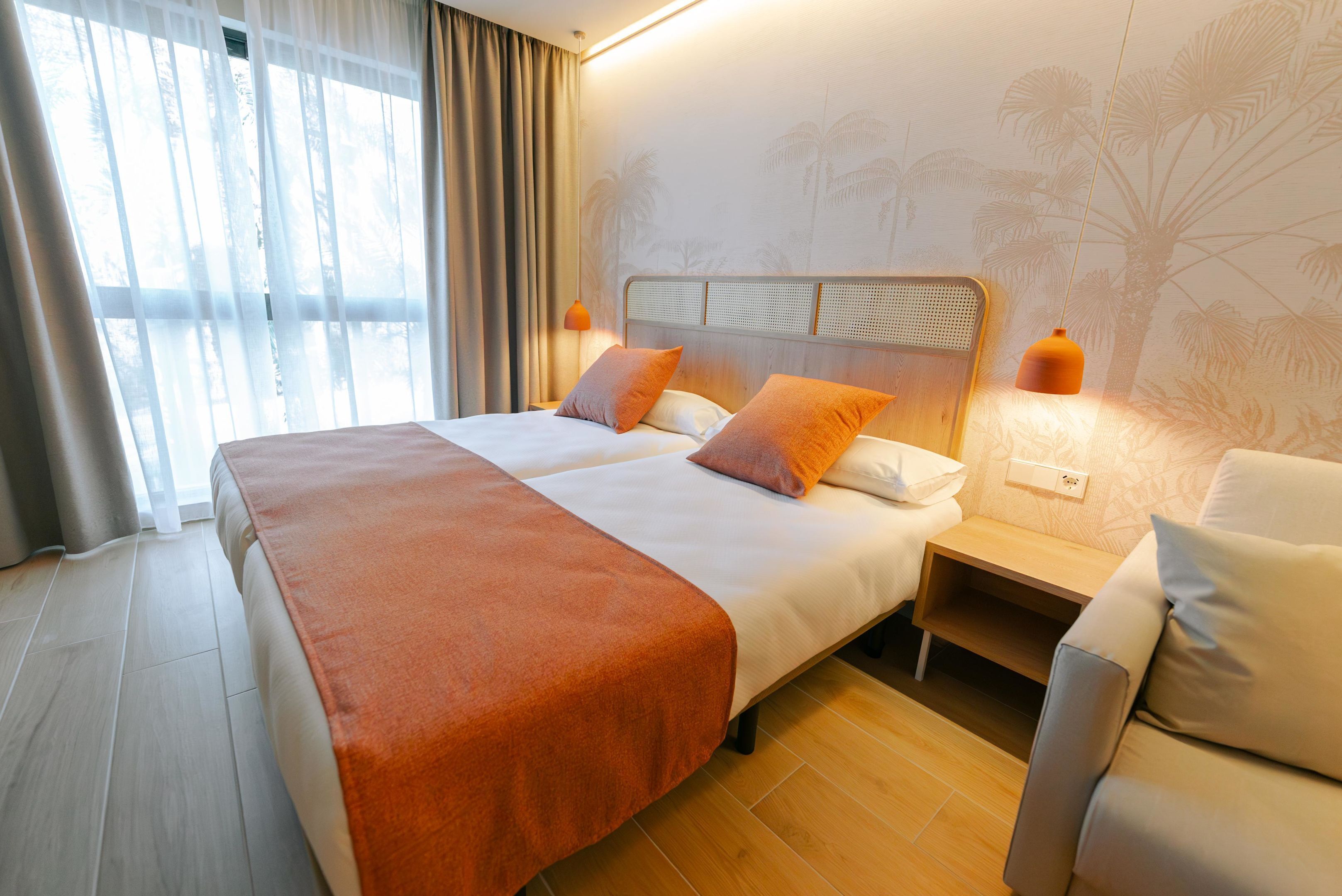 A double bed with two pillows, a nightstand, a lamp, and a wall with palm tree wallpaper.