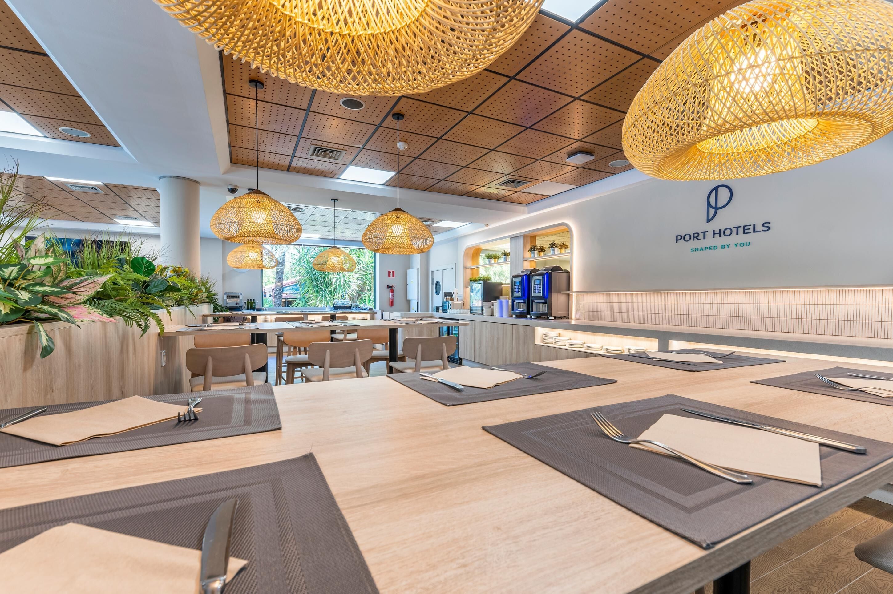 A modern hotel restaurant with wooden tables, woven pendant lights, and a visible logo on the wall.