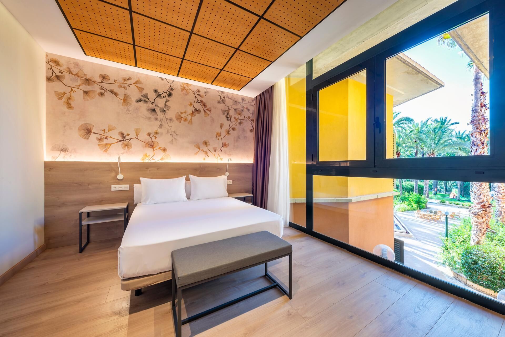 A modern hotel room with a large bed, bedside tables, a bench, a ceiling with a perforated design, and a large window with a view of greenery.
