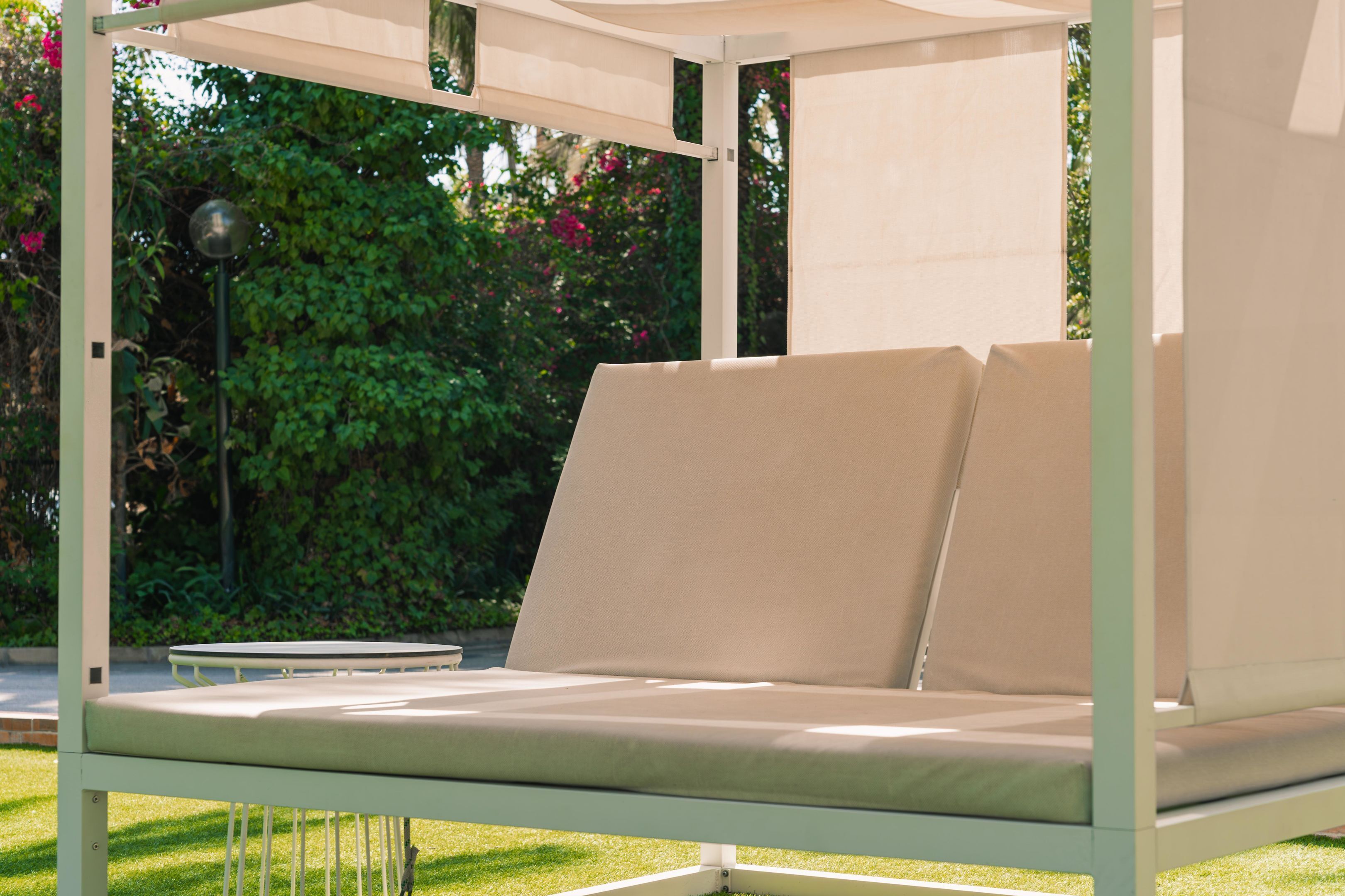 A modern outdoor lounge chair with a light-colored cushion, set against a backdrop of lush greenery and a clear sky.