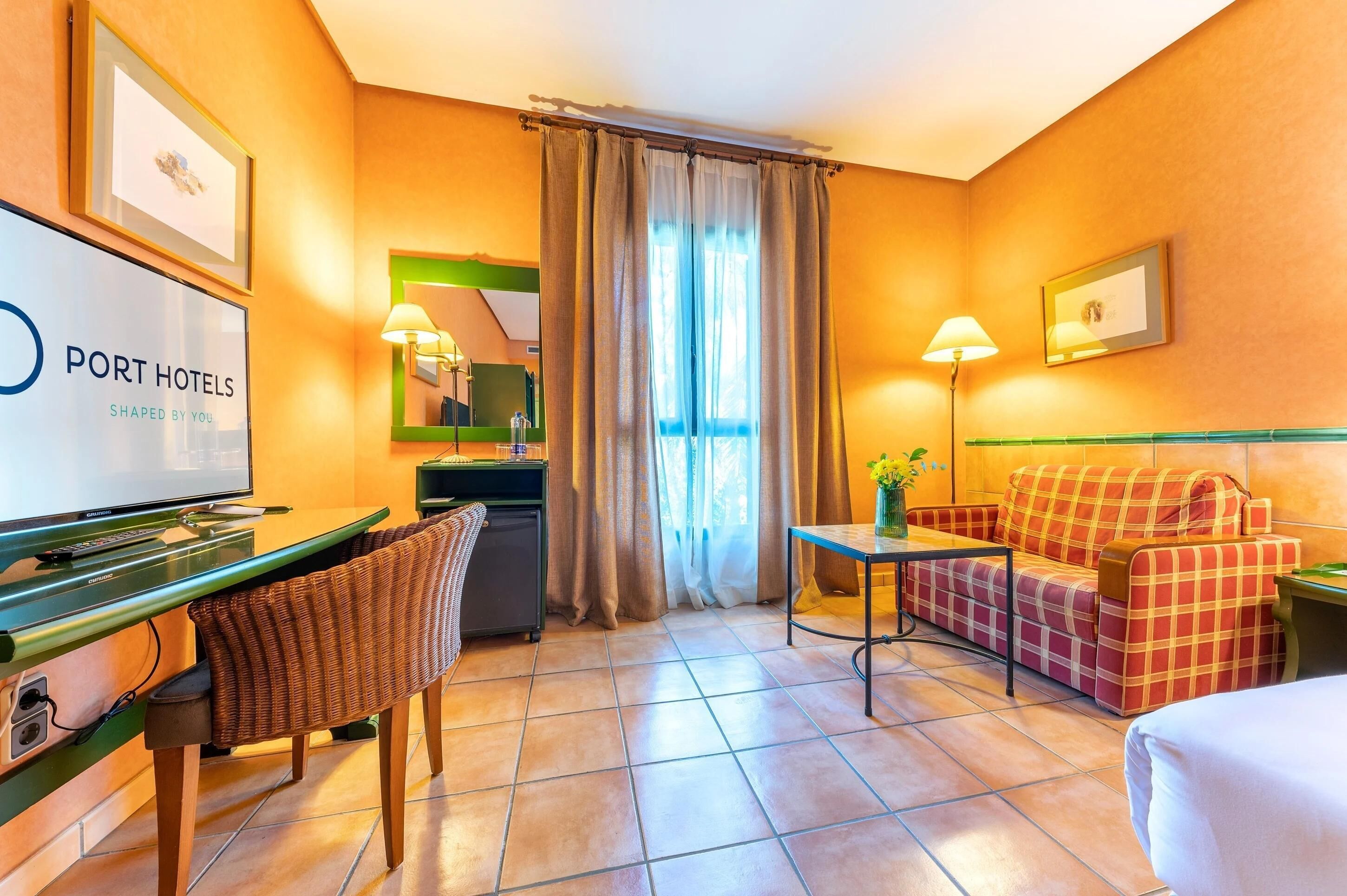 Ein Hotelzimmer mit Bett, einer Couch, einem Schreibtisch mit Computer, einem Sessel und einem Fernseher, der den Namen des Hotels zeigt.