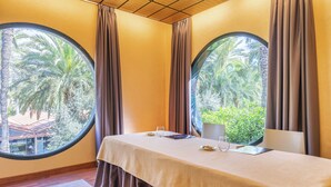Una habitación con una mesa redonda, dos ventanas con cortinas y vistas a palmeras y un edificio.