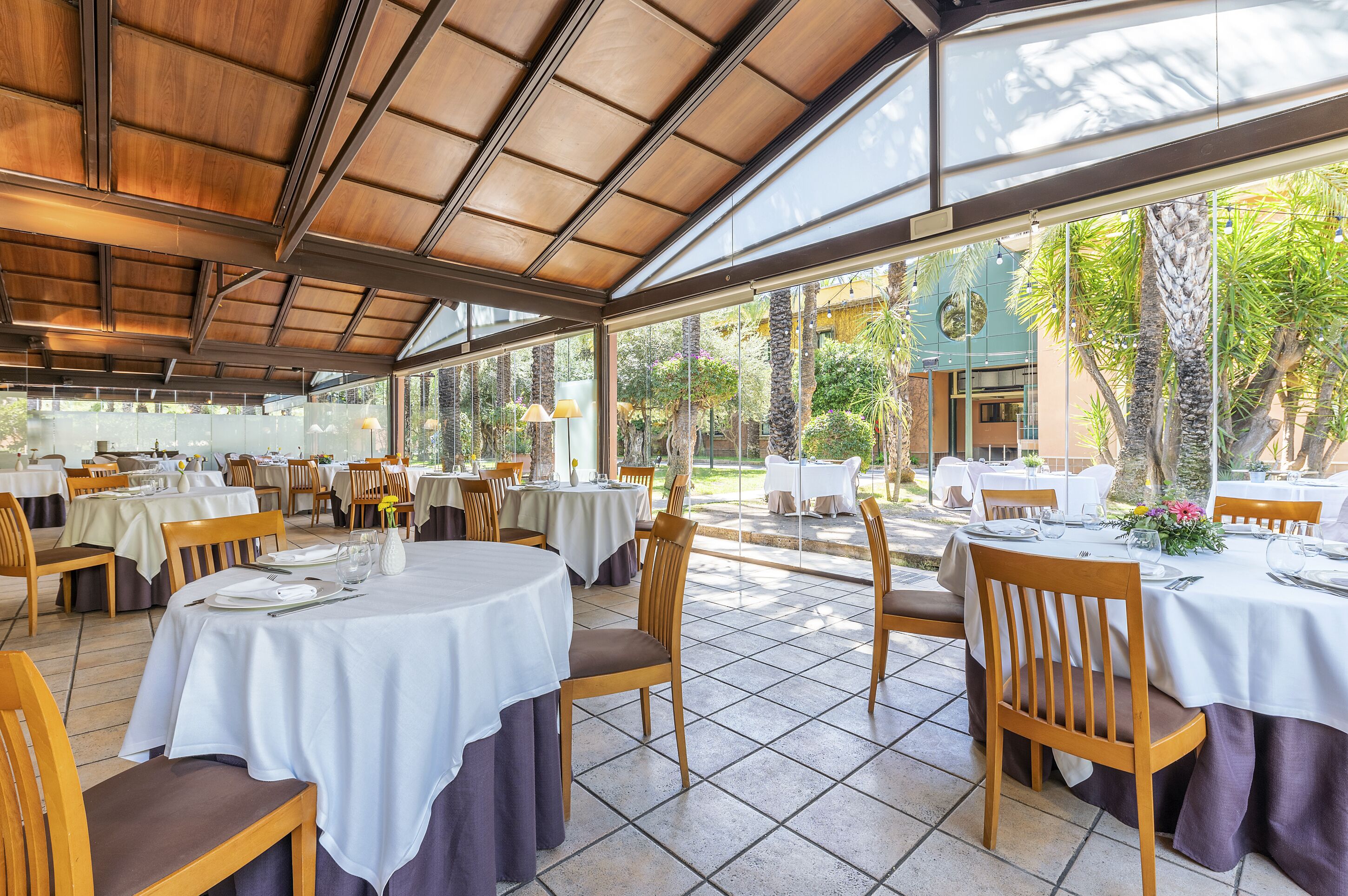 Un restaurante con mesas redondas, sillas de madera y un patio cubierto con paredes de vidrio.