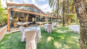 Un área de comedor al aire libre con mesas y sillas, rodeada de palmeras y una estructura de vidrio.