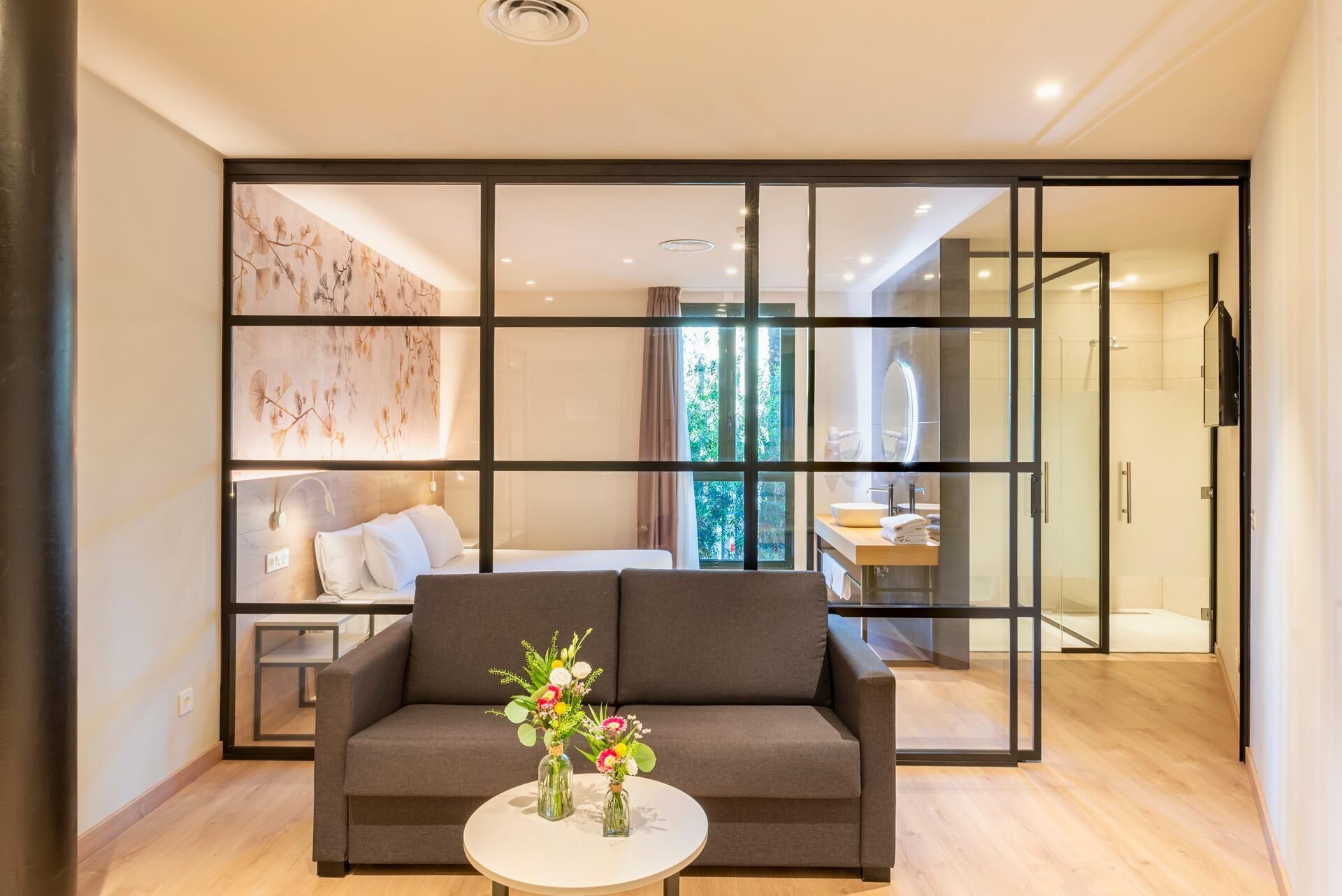 Una sala de estar moderna con un sofá, una mesa de centro con un jarrón de flores y un amplio ventanal corredera que da a un dormitorio con cama y baño.