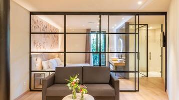 Una sala de estar moderna con un sofá, una mesa de centro con un jarrón de flores y un amplio ventanal corredera que da a un dormitorio con cama y baño.