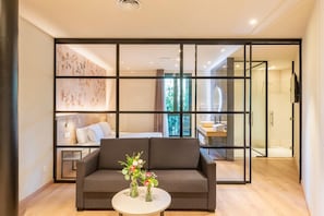 Una sala de estar moderna con un sofá, una mesa de centro con un jarrón de flores y un amplio ventanal corredera que da a un dormitorio con cama y baño.