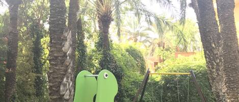 A playground with a green and orange slide, surrounded by palm trees and a grassy area.