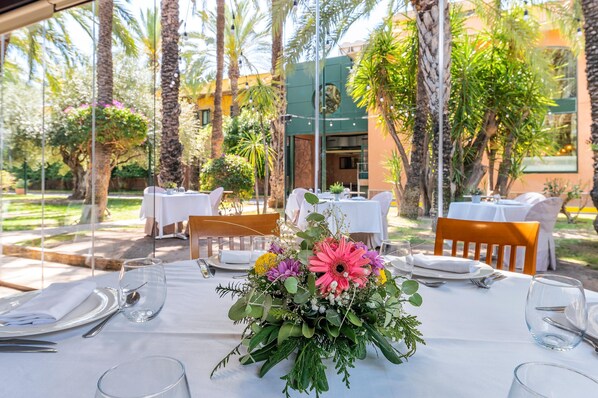 Un área de comedor al aire libre con mesas dispuestas para dos, rodeada de abundante follaje y palmeras.