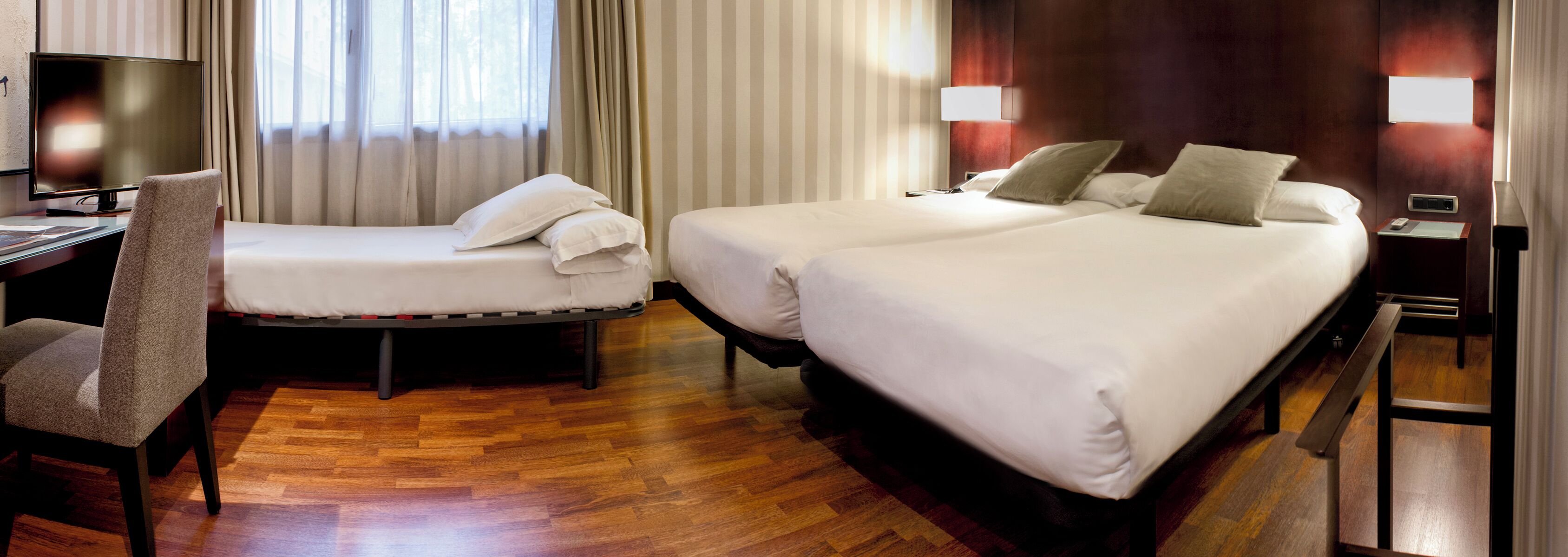 A hotel room with a desk, two beds, a chair, and a TV.