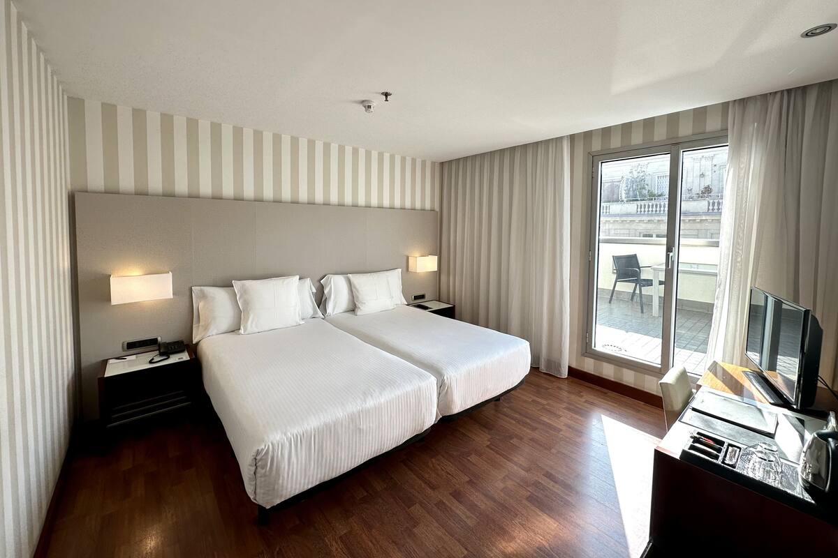 A hotel room with two beds, a desk with a computer, a TV, and a large window with curtains.