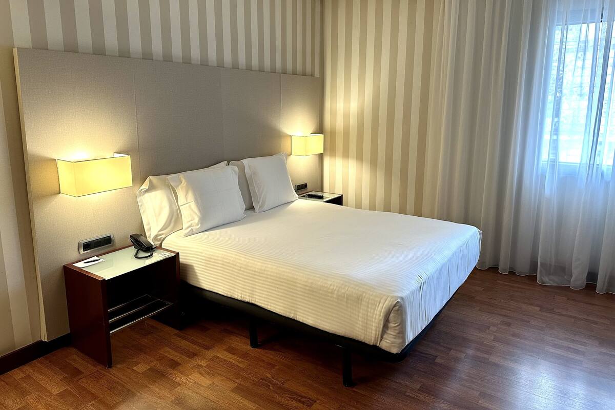 A hotel room with a large bed, two bedside tables, a nightstand, a lamp, and a window with curtains.