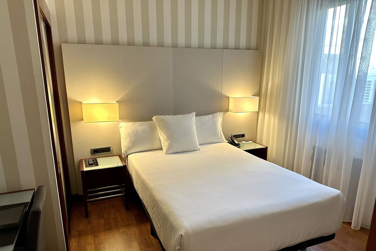 A hotel room with a large bed, two bedside lamps, a nightstand with a phone, and a view of the outdoors through sheer curtains.