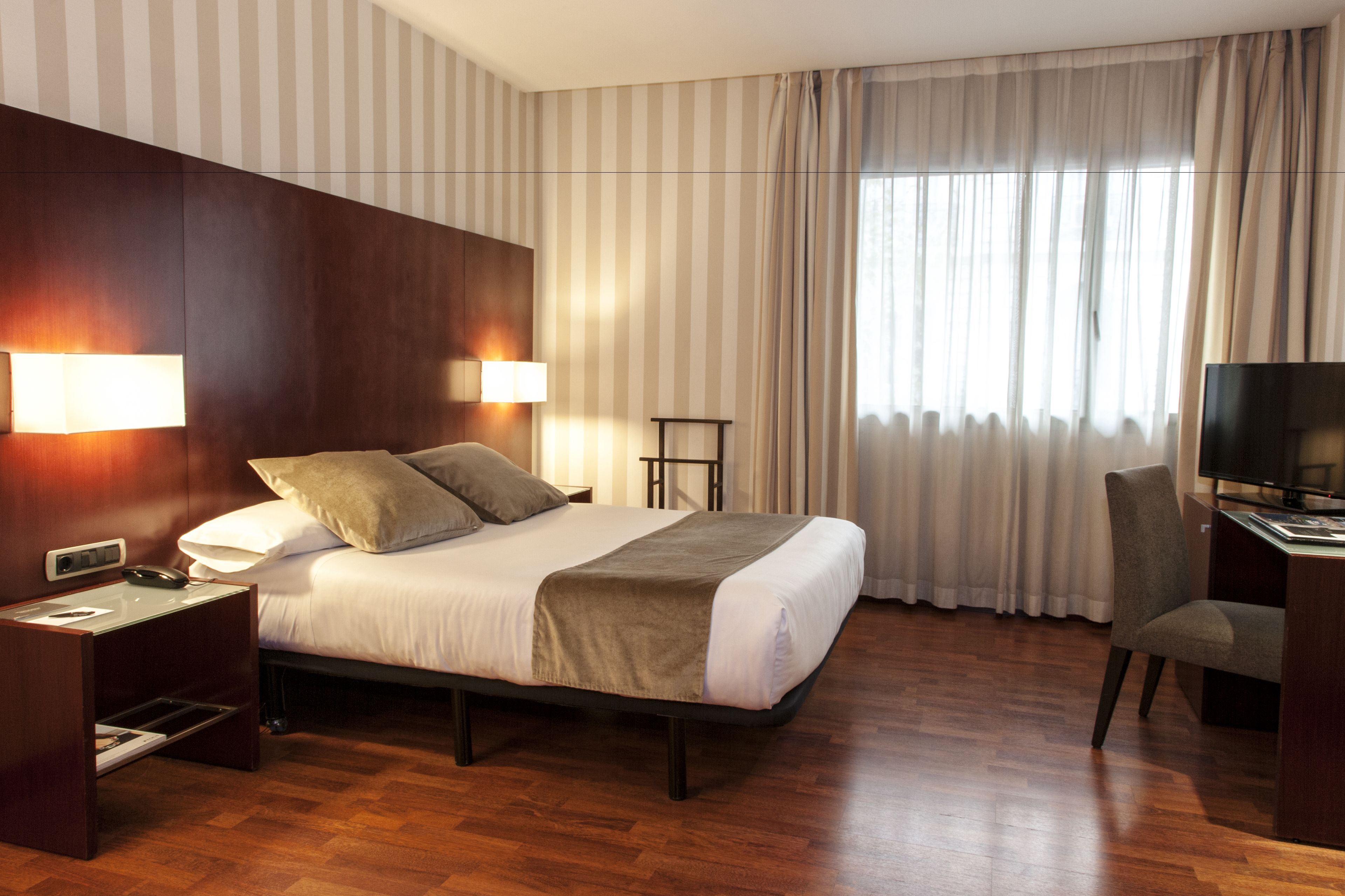 Habitación de hotel con una cama grande, dos lámparas de noche, un escritorio con silla, televisor y ventana con cortinas.