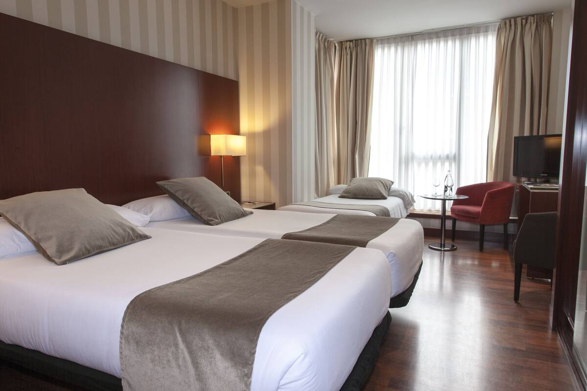 A hotel room with two beds, a desk, a chair, a TV, and a window with curtains.