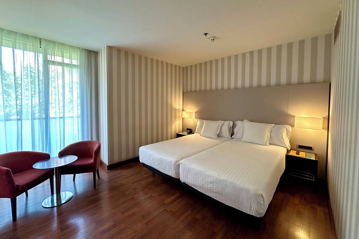 A hotel room with a double bed, two bedside tables with lamps, a small round table with a chair, and a large window with curtains.