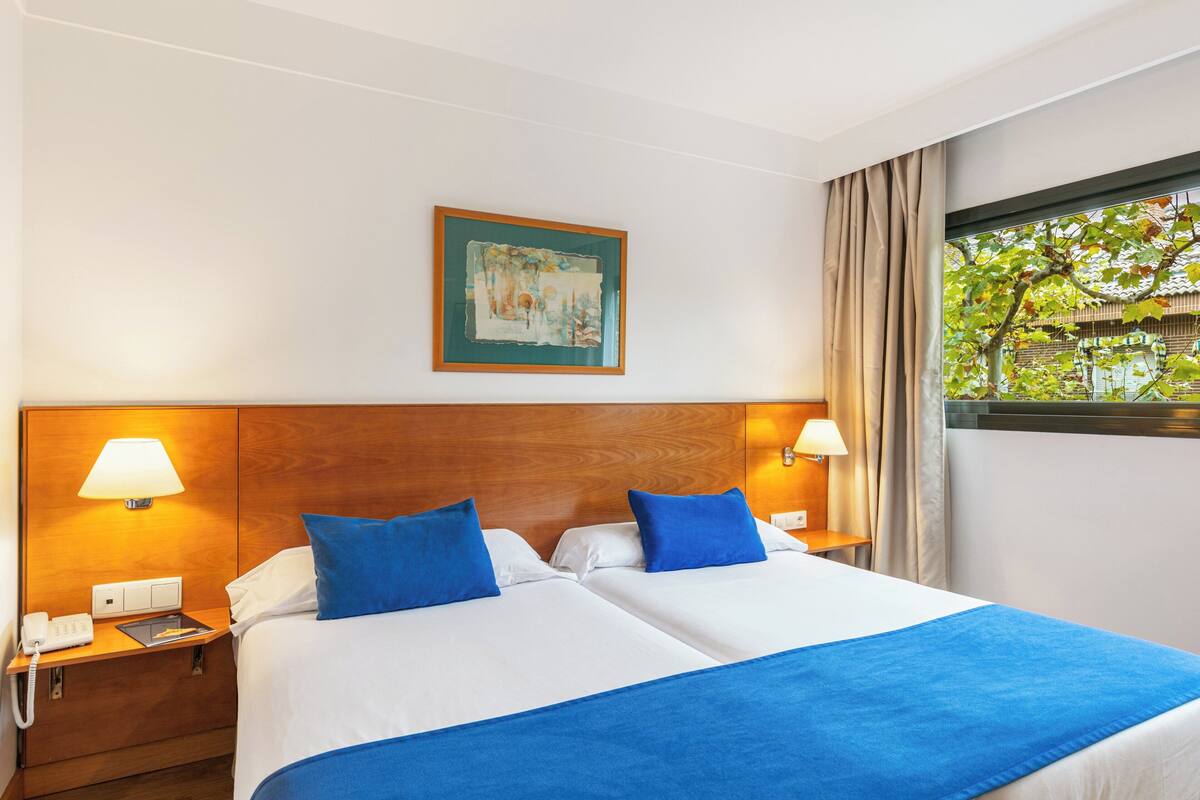 Habitación de hotel con dos camas, cabecero de madera, un cuadro en la pared y ventana con vista a árboles.