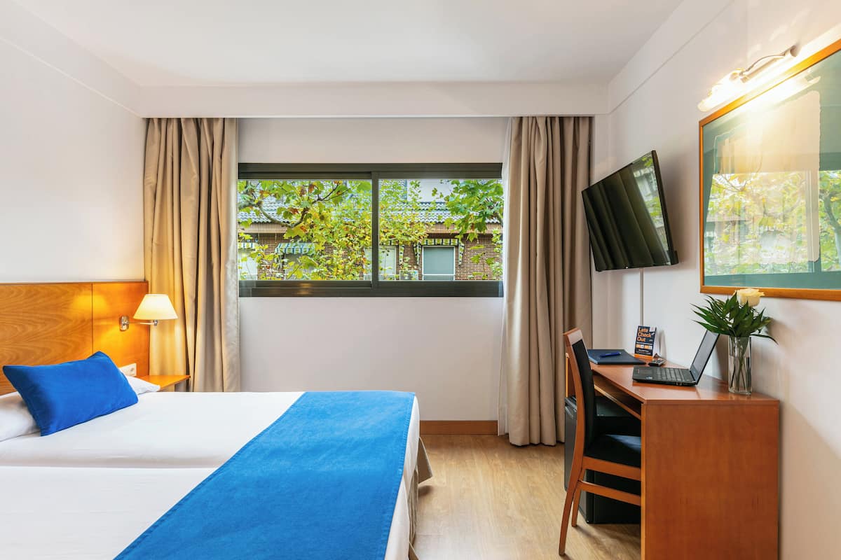 Una habitación de hotel con cama, escritorio, ordenador portátil, televisor y ventana con cortinas.