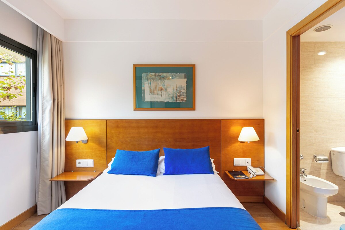 A hotel room with a large bed, two bedside tables, a nightstand, a window with curtains, a wall-mounted painting, and a bathroom visible through an open door.