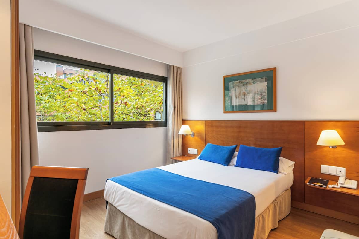 Una habitación de hotel con una cama grande, dos lámparas de noche, una mesita de noche, un ventanal con vistas a árboles y un cuadro en la pared.
