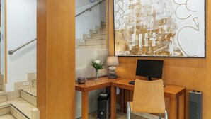 A modern office space with a wooden desk, a computer, and a chair. There is a staircase in the background and a large abstract painting on the wall. - Sercotel de los Reyes (San Sebastian de los Reyes)