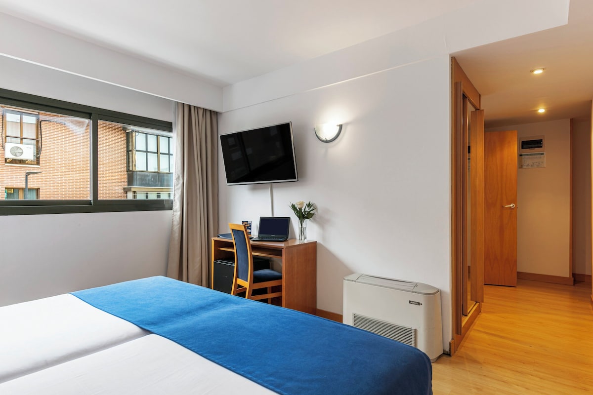 A hotel room with a bed, a desk with a computer, a TV, and a window with curtains.