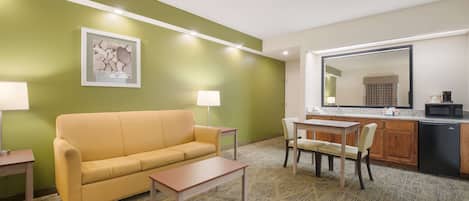 A hotel room with a yellow sofa, a small table, a chair, a kitchen area with a microwave, and a large mirror.
