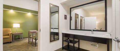 Un baño de hotel con un espejo grande, un lavabo y un inodoro. En la habitación contigua hay una mesita con una lámpara y una silla.