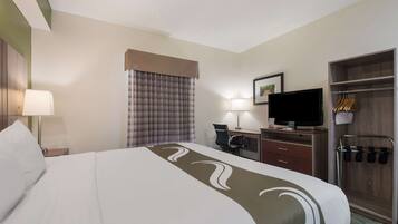 Ein Hotelzimmer mit einem großen Bett, einem Schreibtisch mit Computer, einem Fernseher und einem Kleiderschrank mit Kleidung.