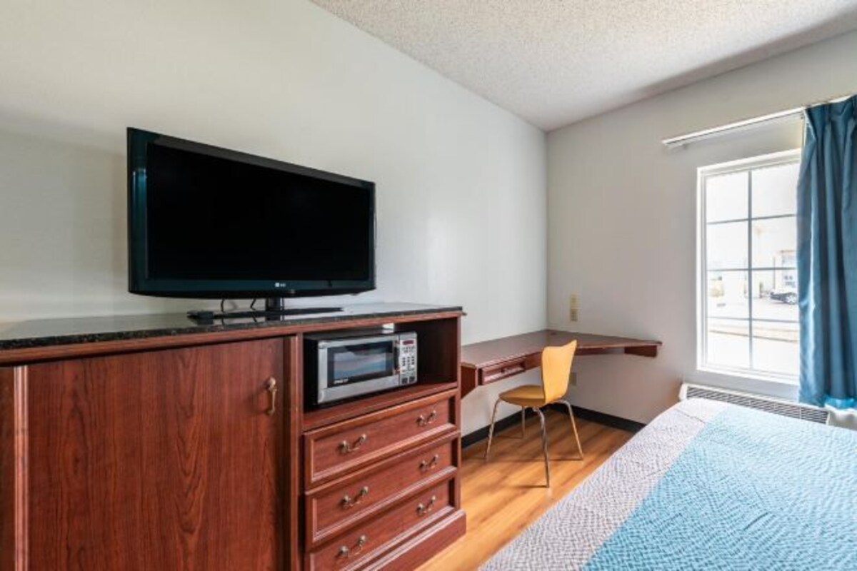 Une chambre d’hôtel équipée d’un meuble en bois, d’un téléviseur à écran plat, d’un four à micro-ondes, d’un bureau, d’une chaise et d’une grande fenêtre avec des rideaux.