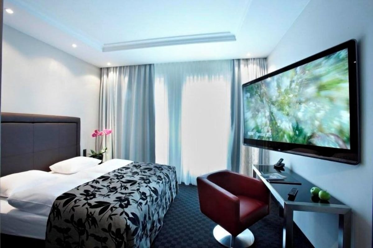 Una habitación de hotel con cama, escritorio, televisor de pantalla plana, una silla roja y lámpara de escritorio.