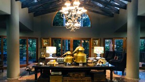 Une pièce spacieuse, ornée d’un grand lustre, avec une table en bois sur laquelle sont disposés divers objets, et offrant une vue sur l’extérieur grâce à de grandes fenêtres.
