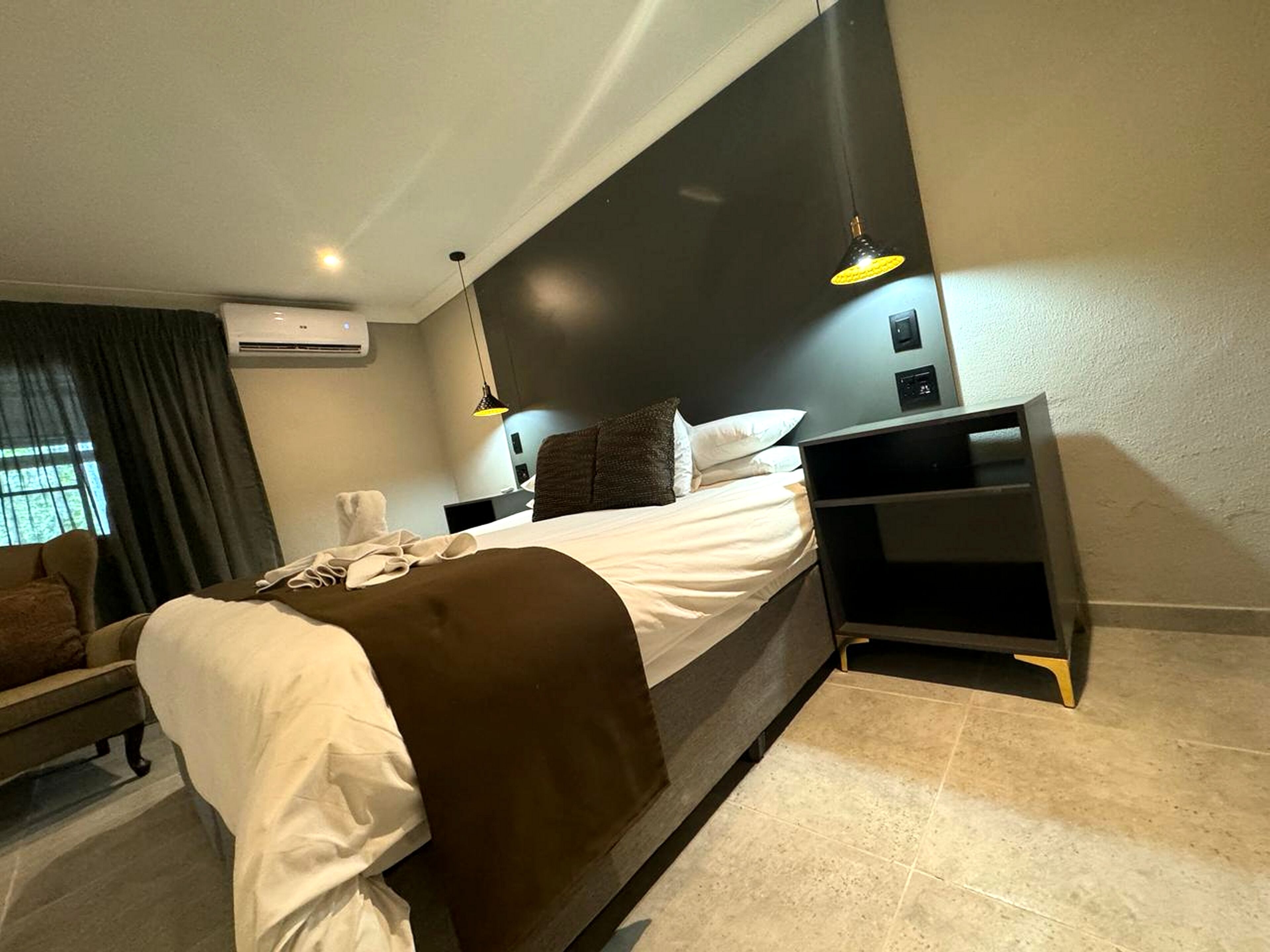 Una habitación de hotel moderna con una cama grande, una mesita de noche, un equipo de aire acondicionado y un ventanal con cortinas.