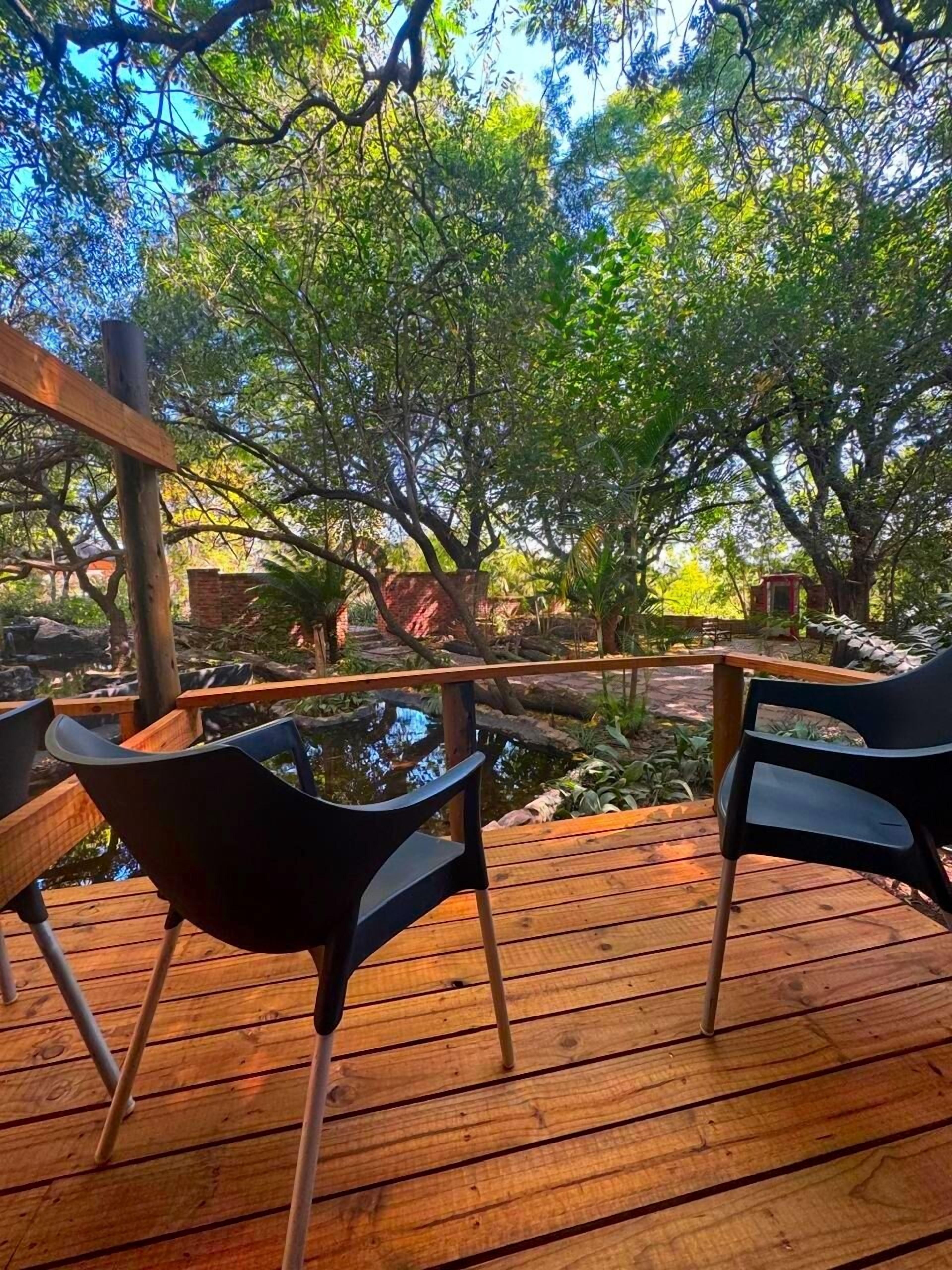 Una terraza de madera con dos sillas negras orientadas hacia un estanque, rodeado de exuberante vegetación y árboles.