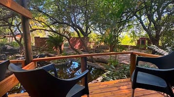 Una terraza de madera con dos sillas negras orientadas hacia un estanque, rodeado de exuberante vegetación y árboles.