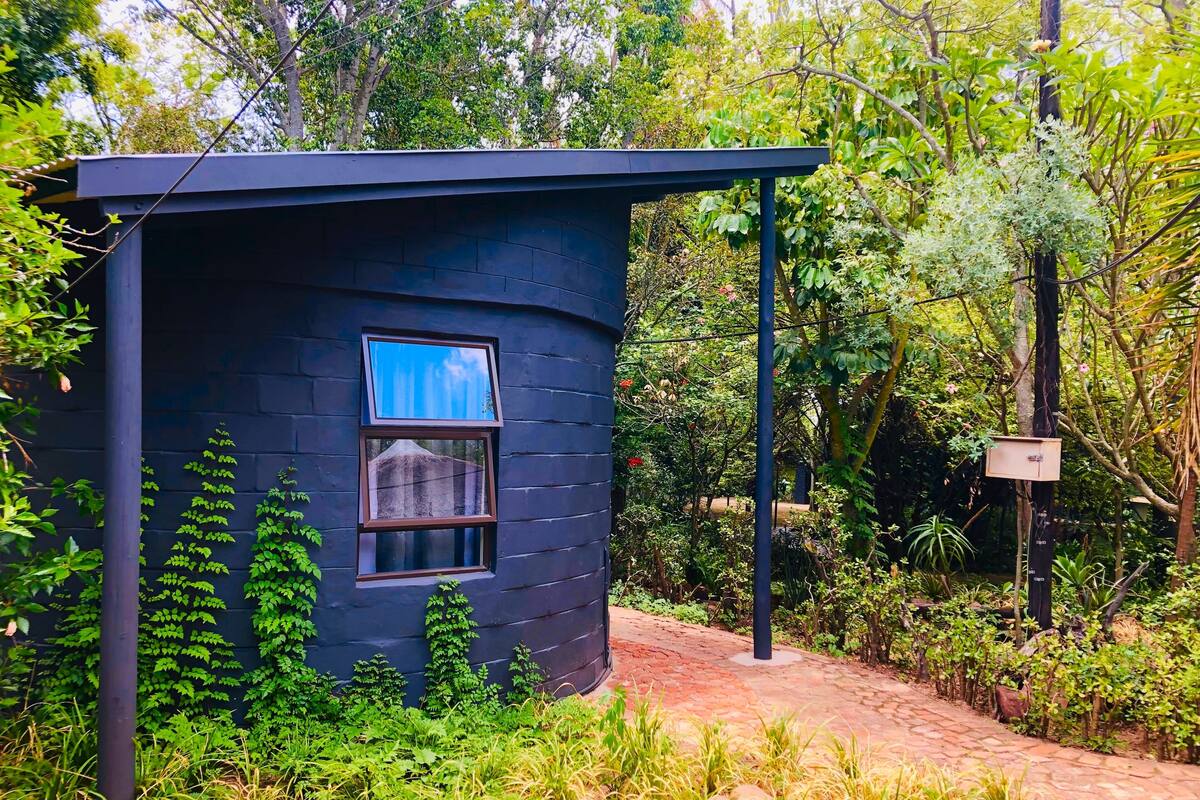 A small, dark-colored cabin with a window, surrounded by lush greenery and a brick pathway.