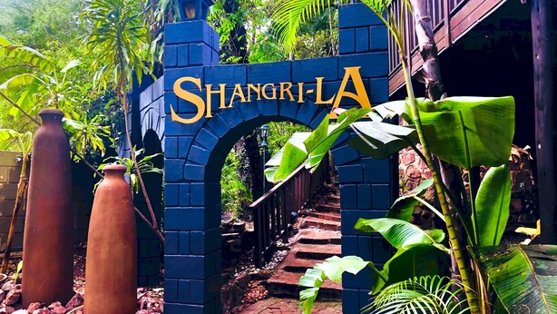Un arc en briques bleues surmonté du mot « SHANGRI-LA » en lettres dorées, qui mène à un sentier bordé de plantes tropicales et doté d’un sol en briques.