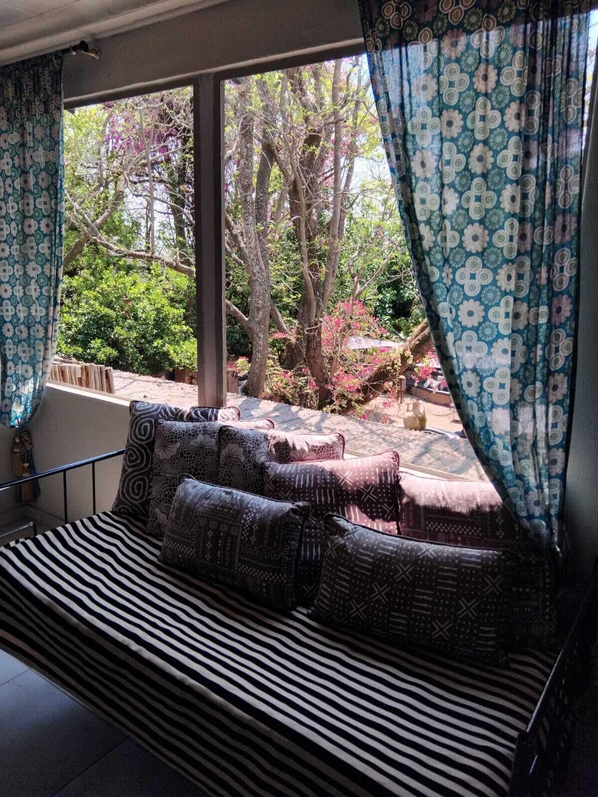 A room with a large window, a sofa with patterned cushions, and a view of trees and greenery outside.