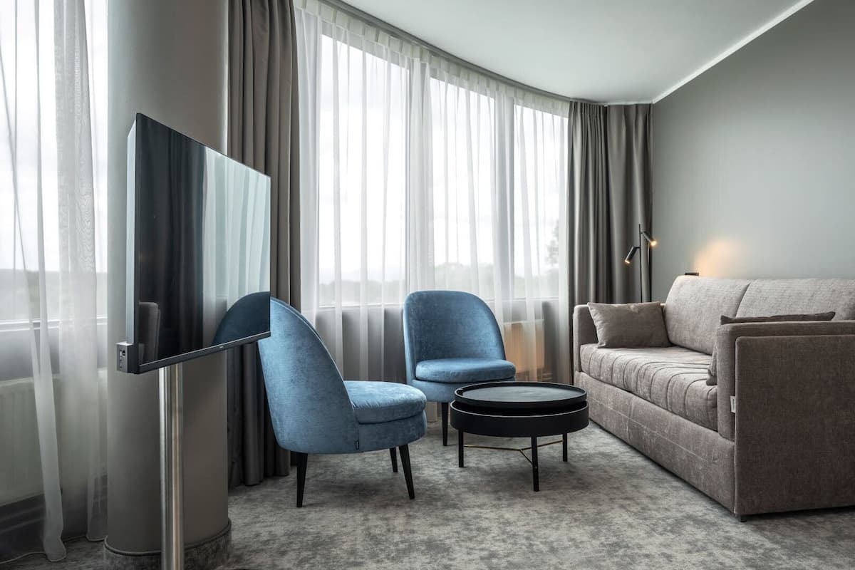 A modern hotel room with a grey sofa, a blue armchair, a small round coffee table, and a flat-screen TV mounted on the wall.