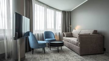 Una habitación de hotel moderna con un sofá gris, un sillón azul, una pequeña mesa redonda de centro y un televisor de pantalla plana montado en la pared.