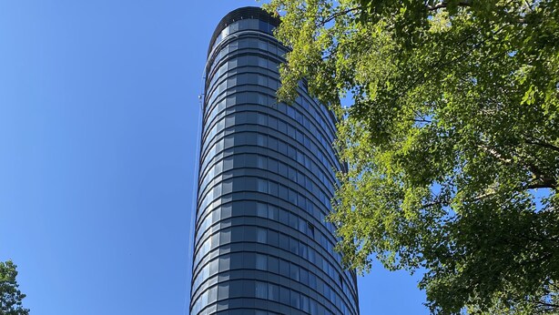 A modern cylindrical building with a glass facade, surrounded by greenery and a clear blue sky.